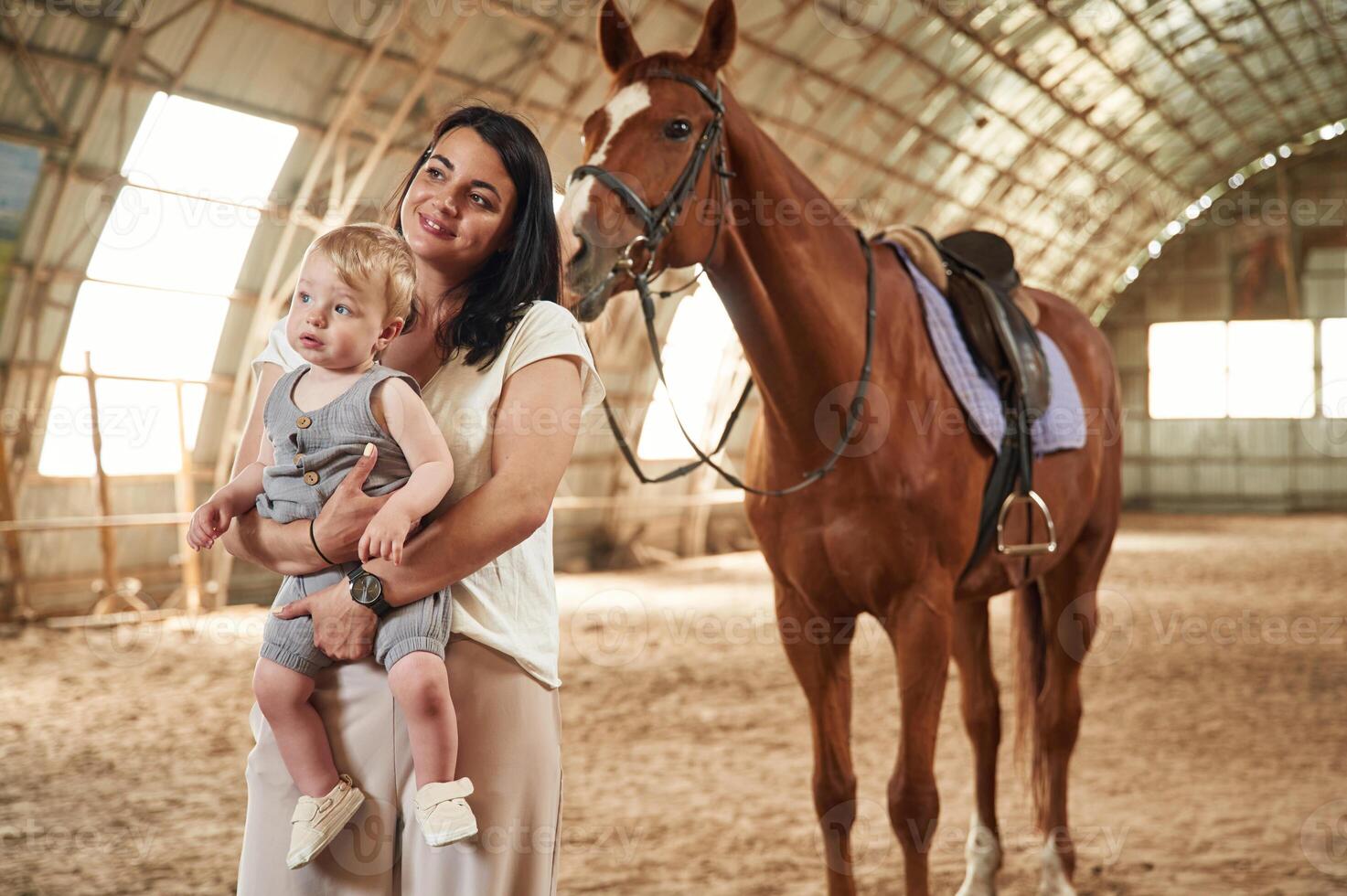 Holding, kid, in, hands. Gorgeous woman with her little baby son is with horse indoors photo