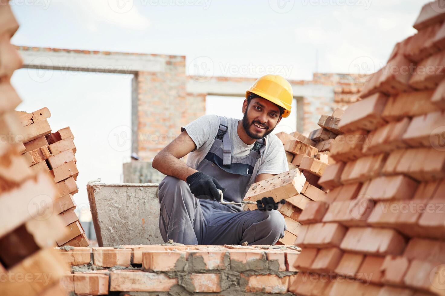 Sitting, and holding brick and little showel. Handsome Indian man is on the construction site photo