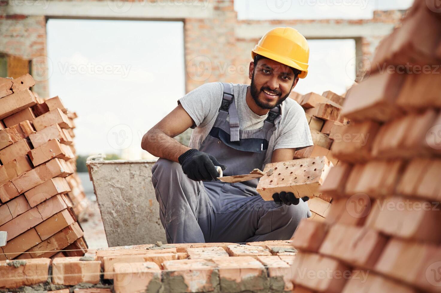 Sitting and, holding brick and little showel. Handsome Indian man is on the construction site photo