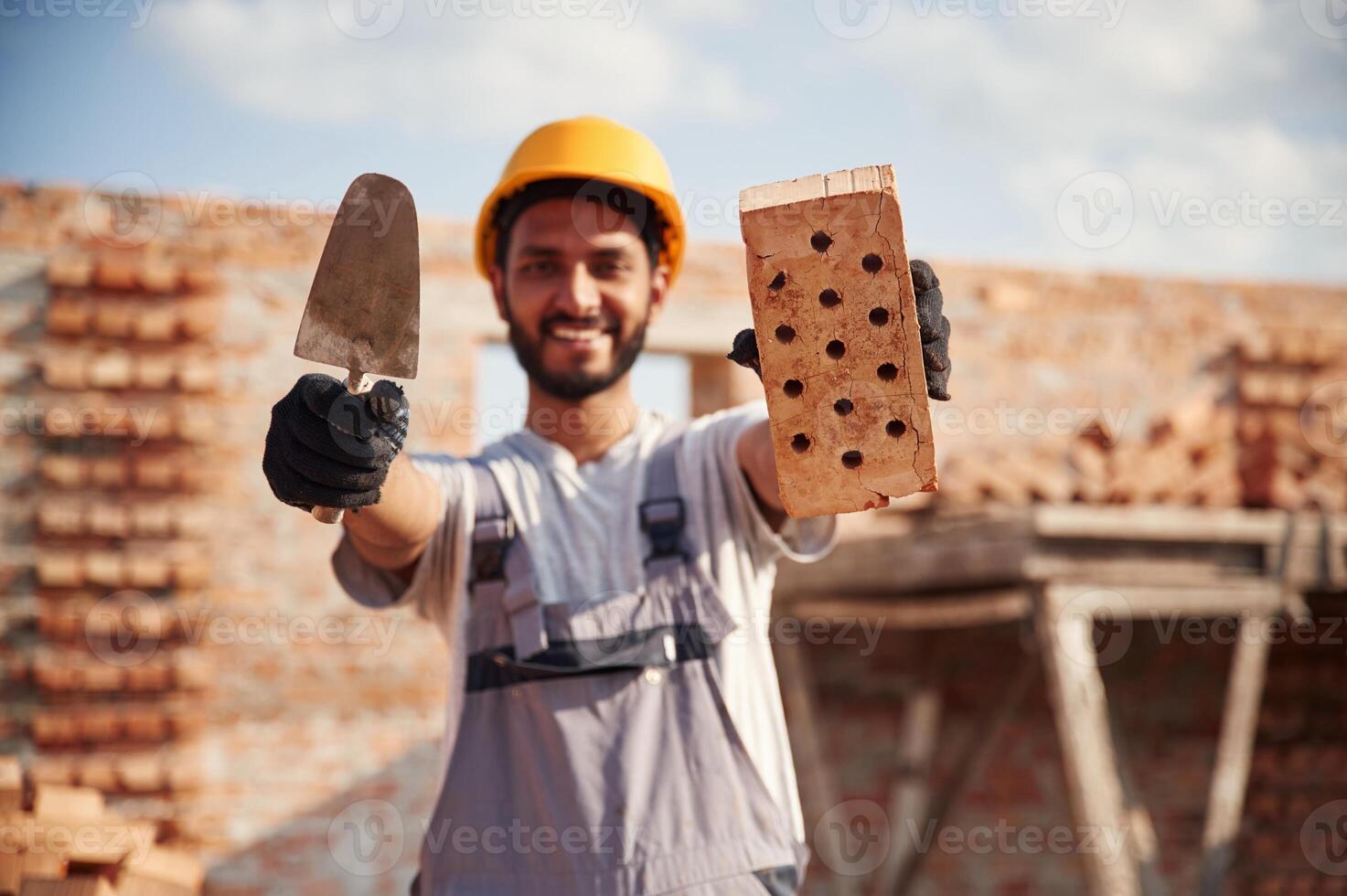 Showing and holding, little ,shovel, and brick. Handsome Indian man is on the construction site photo
