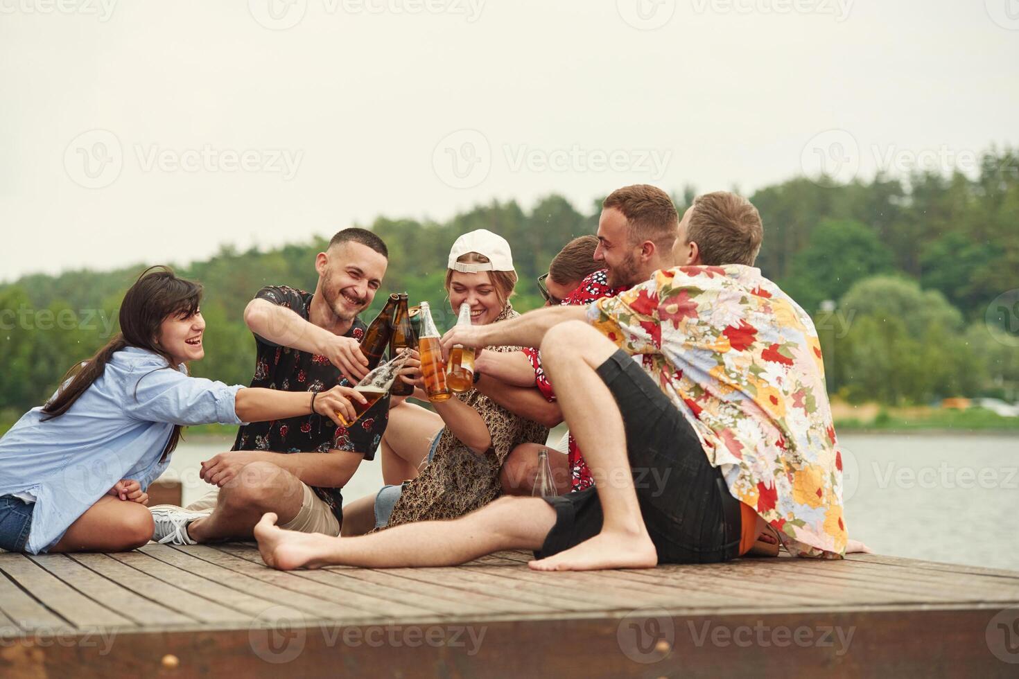 On the wooden floor. Group, of ,friends are sitting near the lake with beer in hands photo