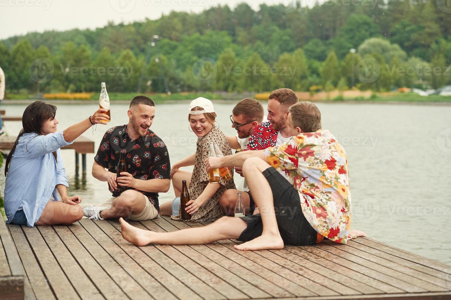 On the, wooden floor. Group of friends are sitting near the lake with beer in hands photo
