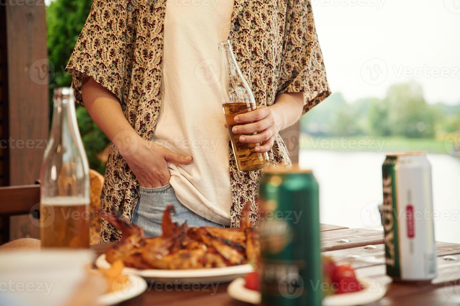 Close up view of woman that is standing by table that is with food, holding beer bottle photo