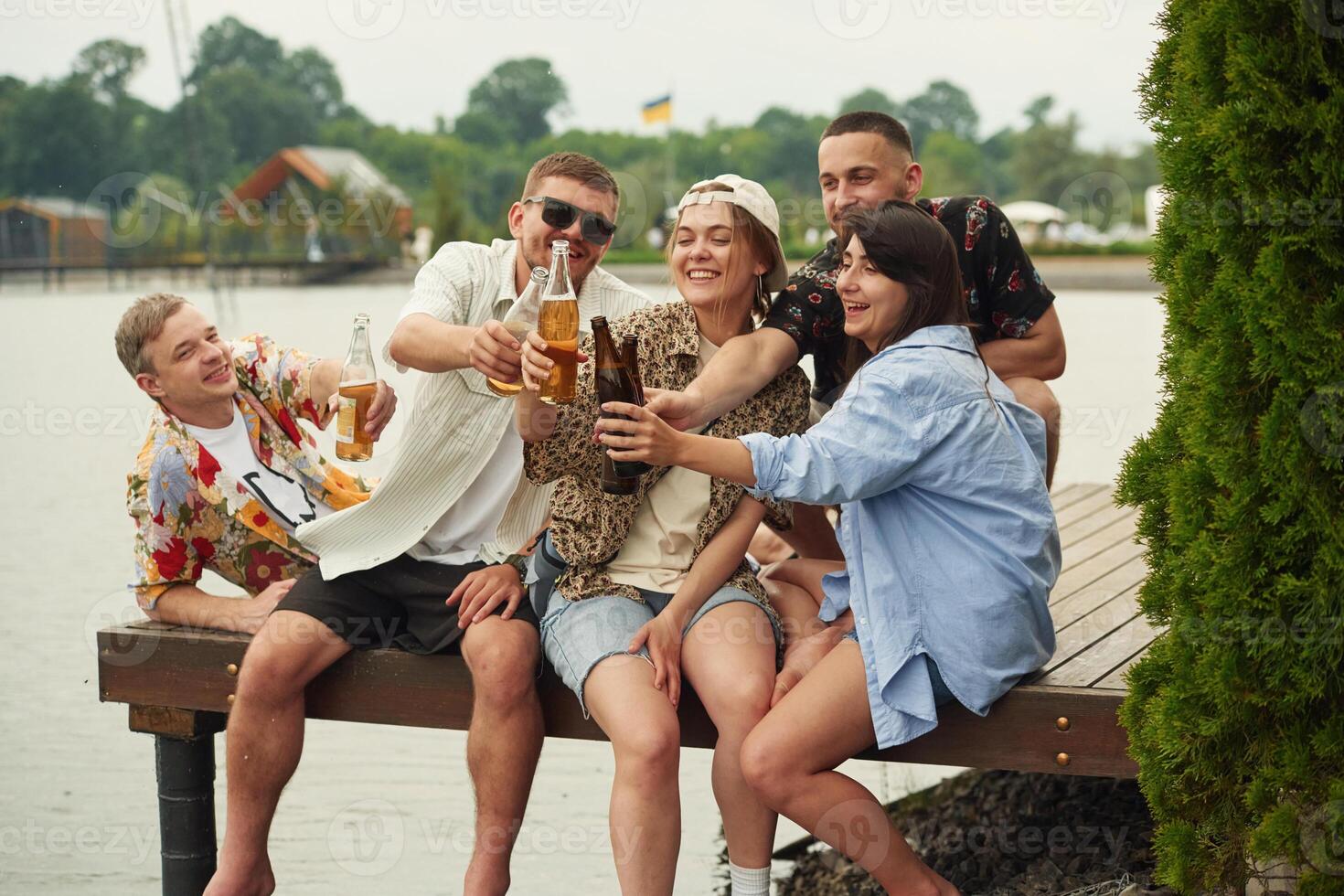 Celebrating,, having fun. Group of friends are sitting near the lake with beer in hands photo