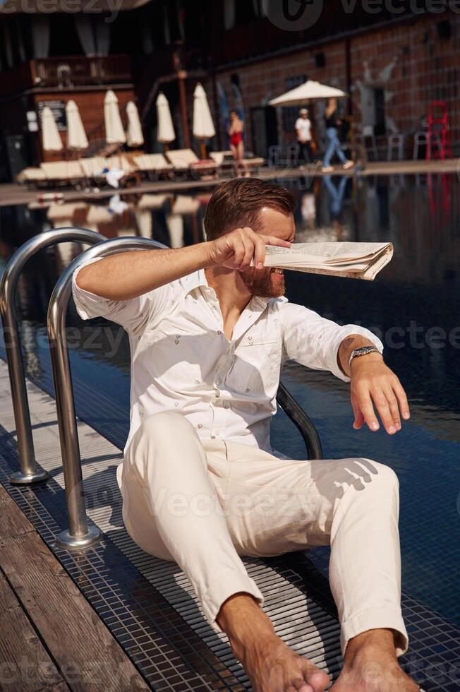Covering himself by newspaper from bright, sunlight. Handsome man in formal clothes is in the pool photo
