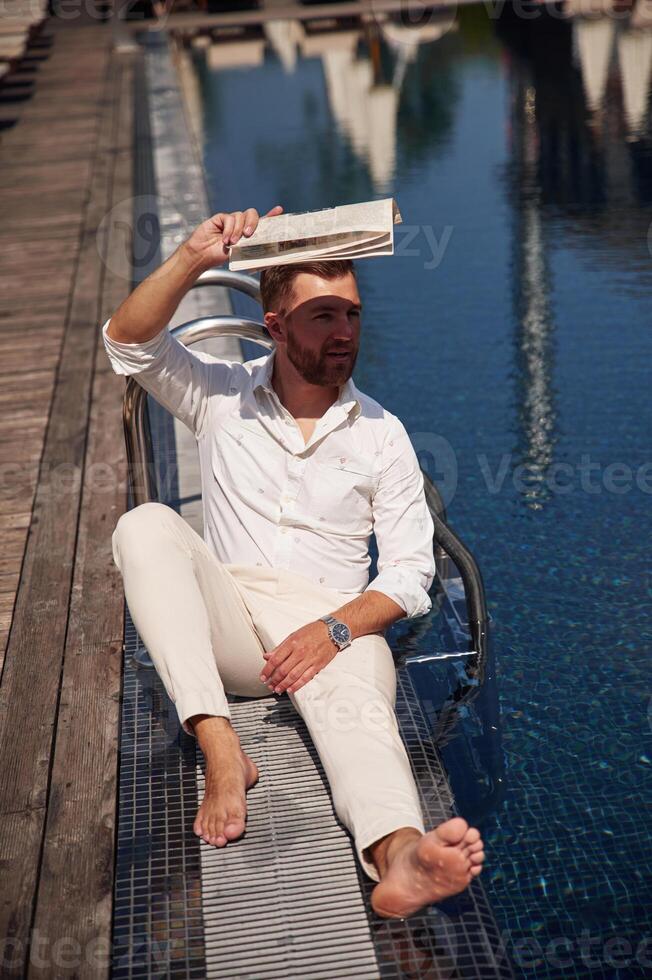 Covering himself, by newspaper from bright sunlight. Handsome man in formal clothes is in the pool photo