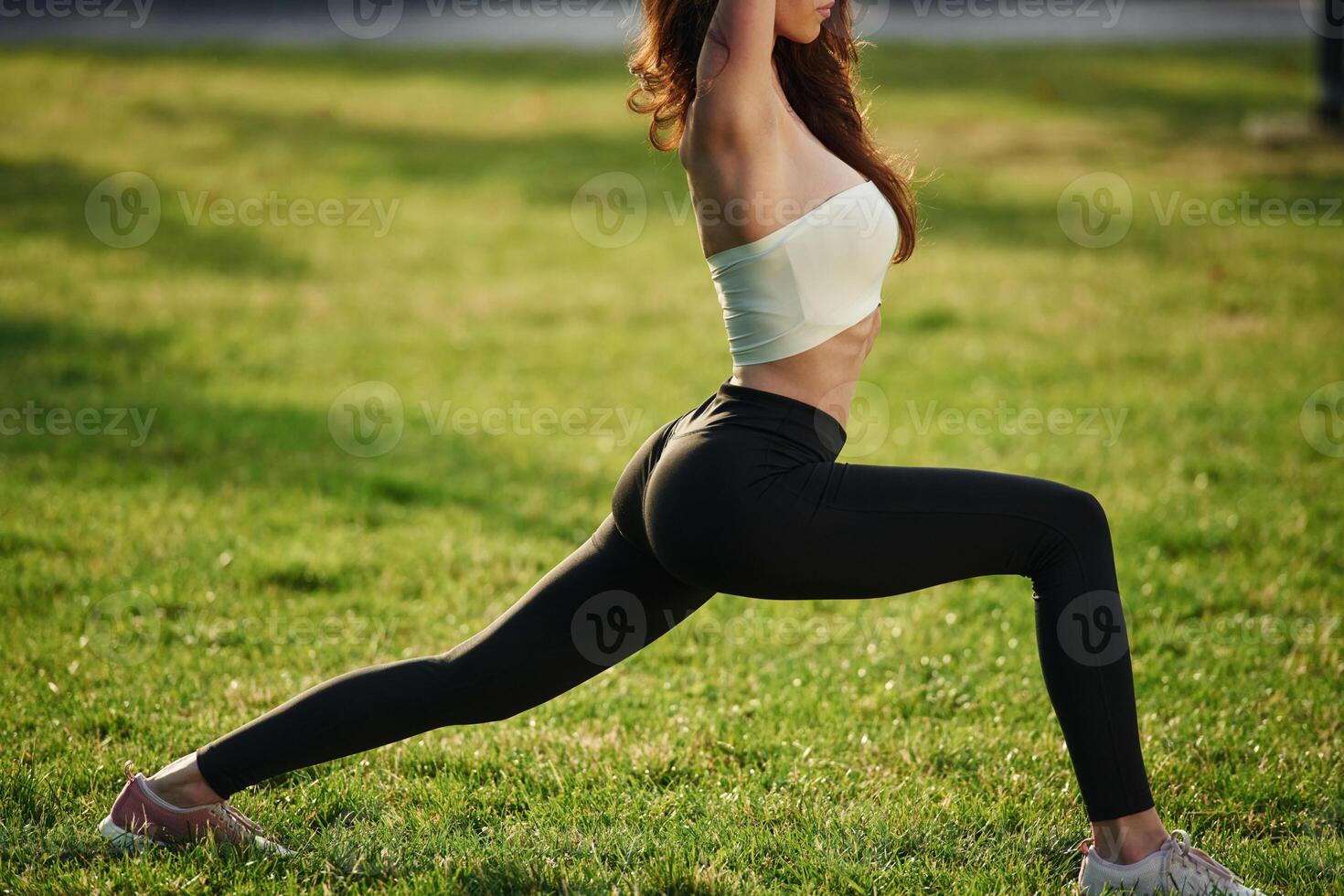 On green grass, stretching the legs. Beautiful young woman in sportive clothes is in the park at sunny daytime photo