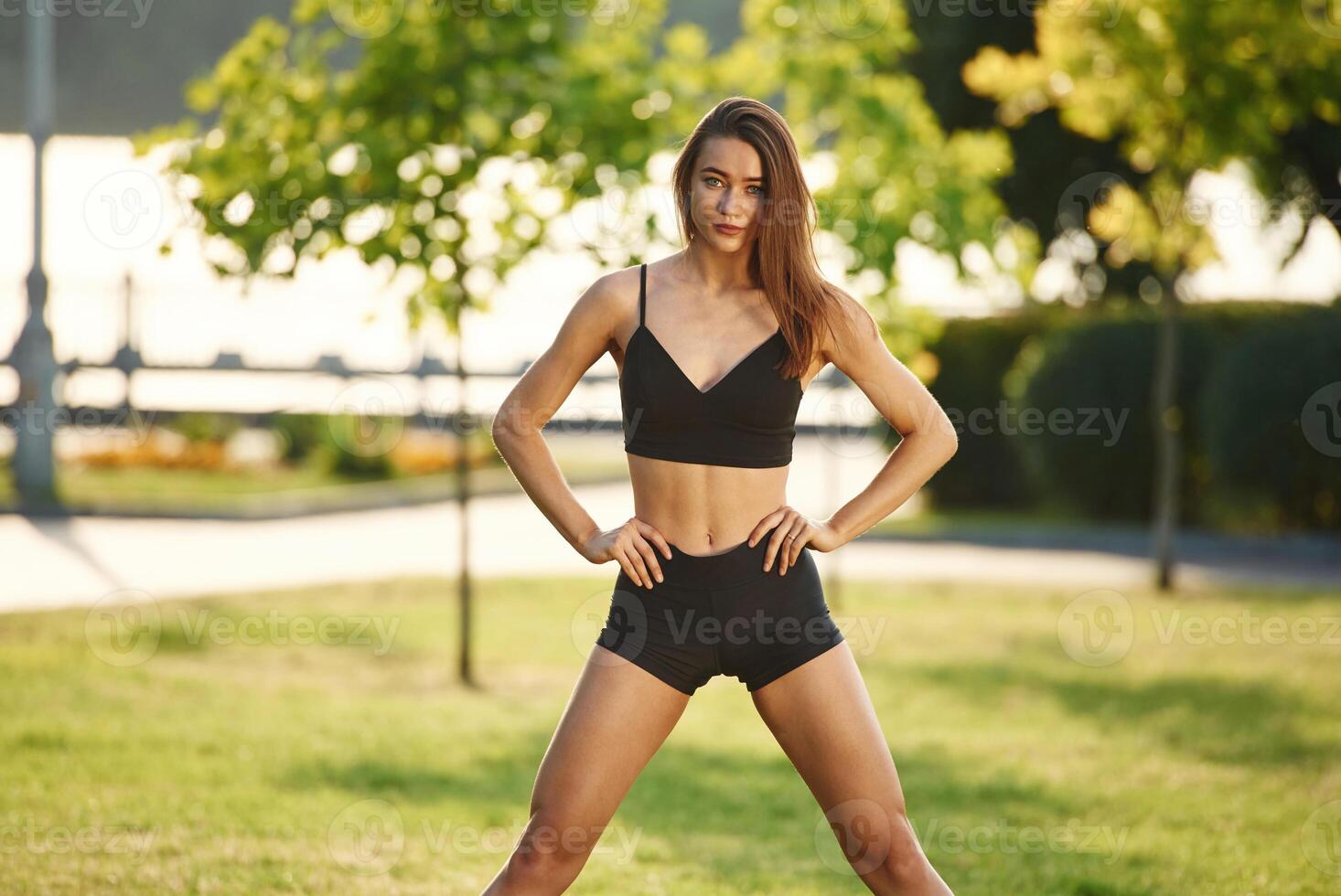 Hands on the waist, standing. Beautiful young woman in sportive clothes is in the park at sunny daytime photo