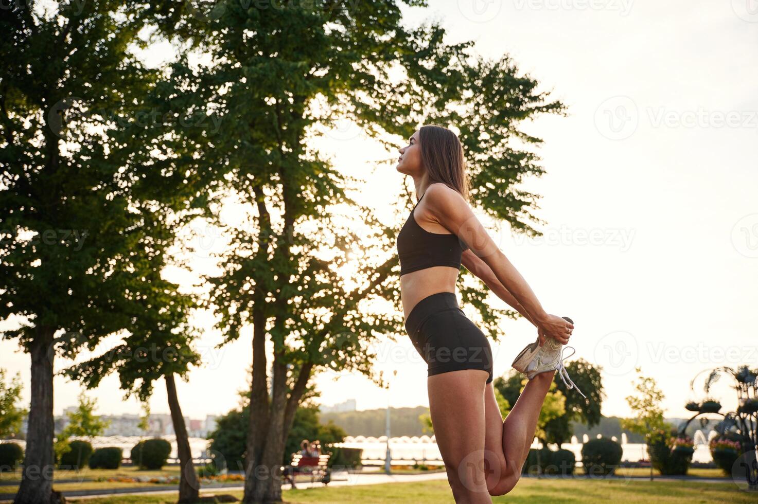 Exercises, against tree and sky... Beautiful young woman in sportive clothes is in the park at sunny daytime photo