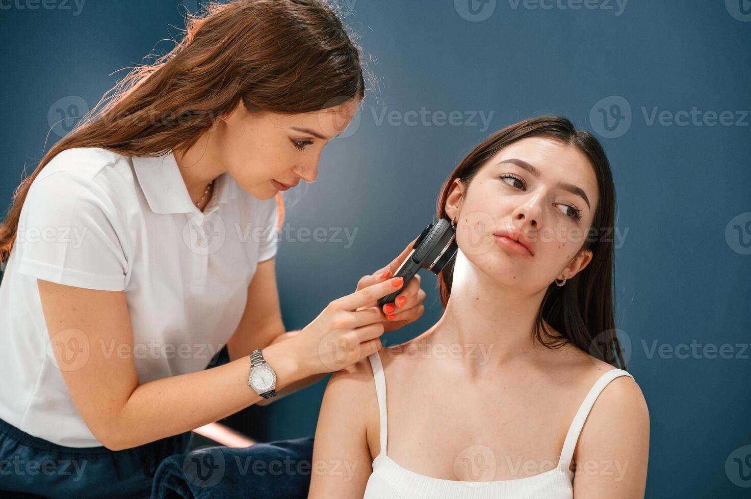 Against blue wall. Beauty salon worker is examining skin of the woman by using a device with light and lens photo