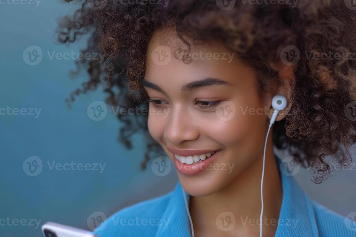 Young Black woman with curly hair listens to music on her smartphone, smiling brightly against a blue backdrop. photo