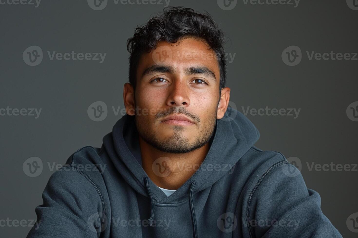 Young Hispanic man with curly hair, wearing a hoodie, gazing confidently at the camera against a muted backdrop. photo