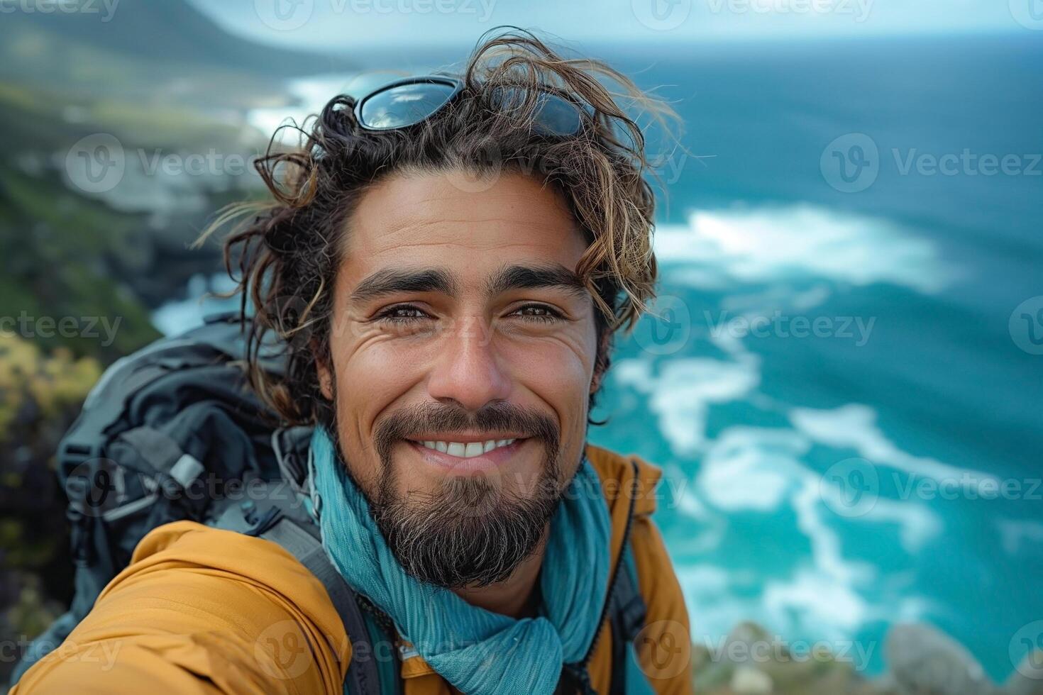 Smiling young man with a beard and wavy hair, wearing hiking gear, enjoying a coastal view. photo