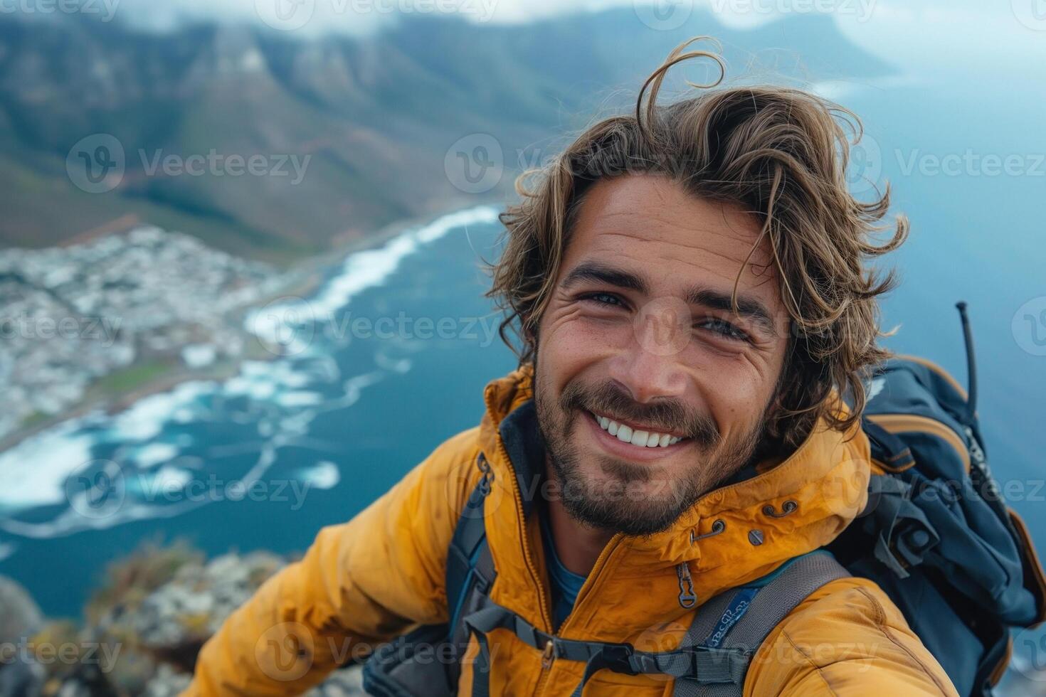 A young Caucasian male adventurer smiles for a selfie atop a mountain, showcasing a rugged landscape in the background. photo