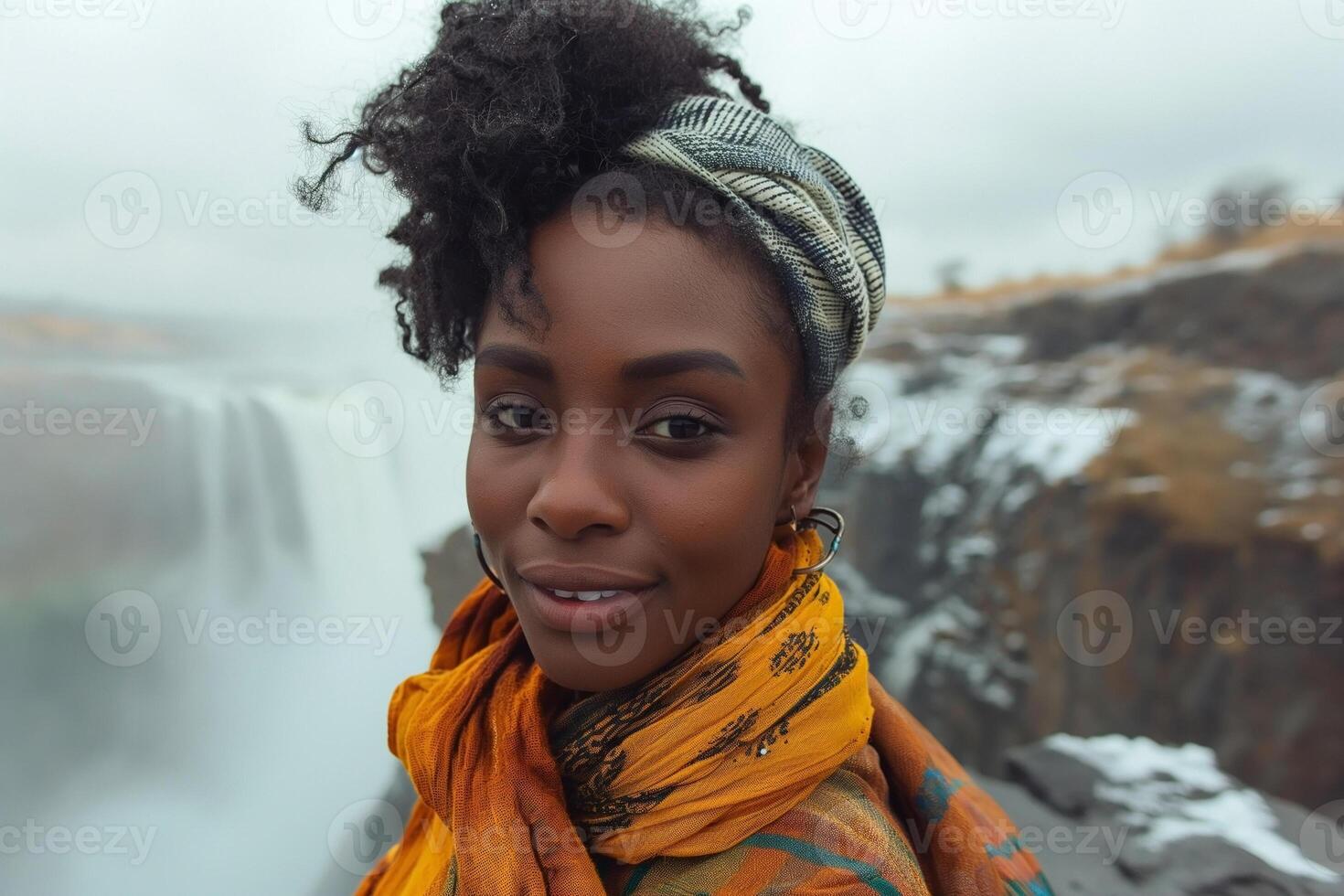 A young Black woman with natural hair, smiling against the backdrop of a stunning waterfall, wearing vibrant colors and a head wrap. photo