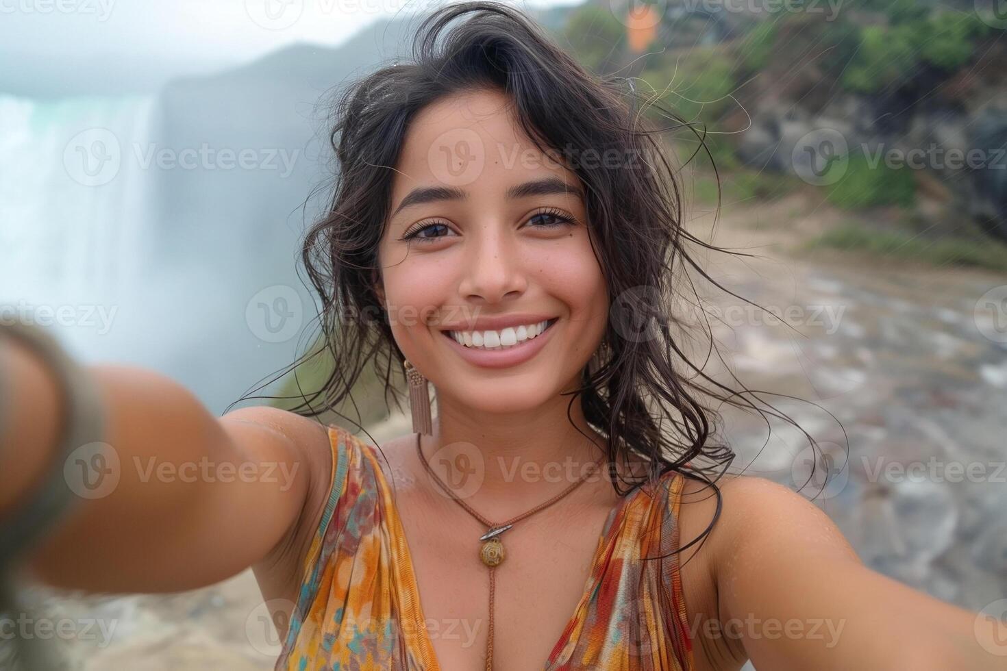A joyful young woman with wavy hair captures a cheerful selfie near cascading waterfalls, radiating natural beauty and happiness. photo