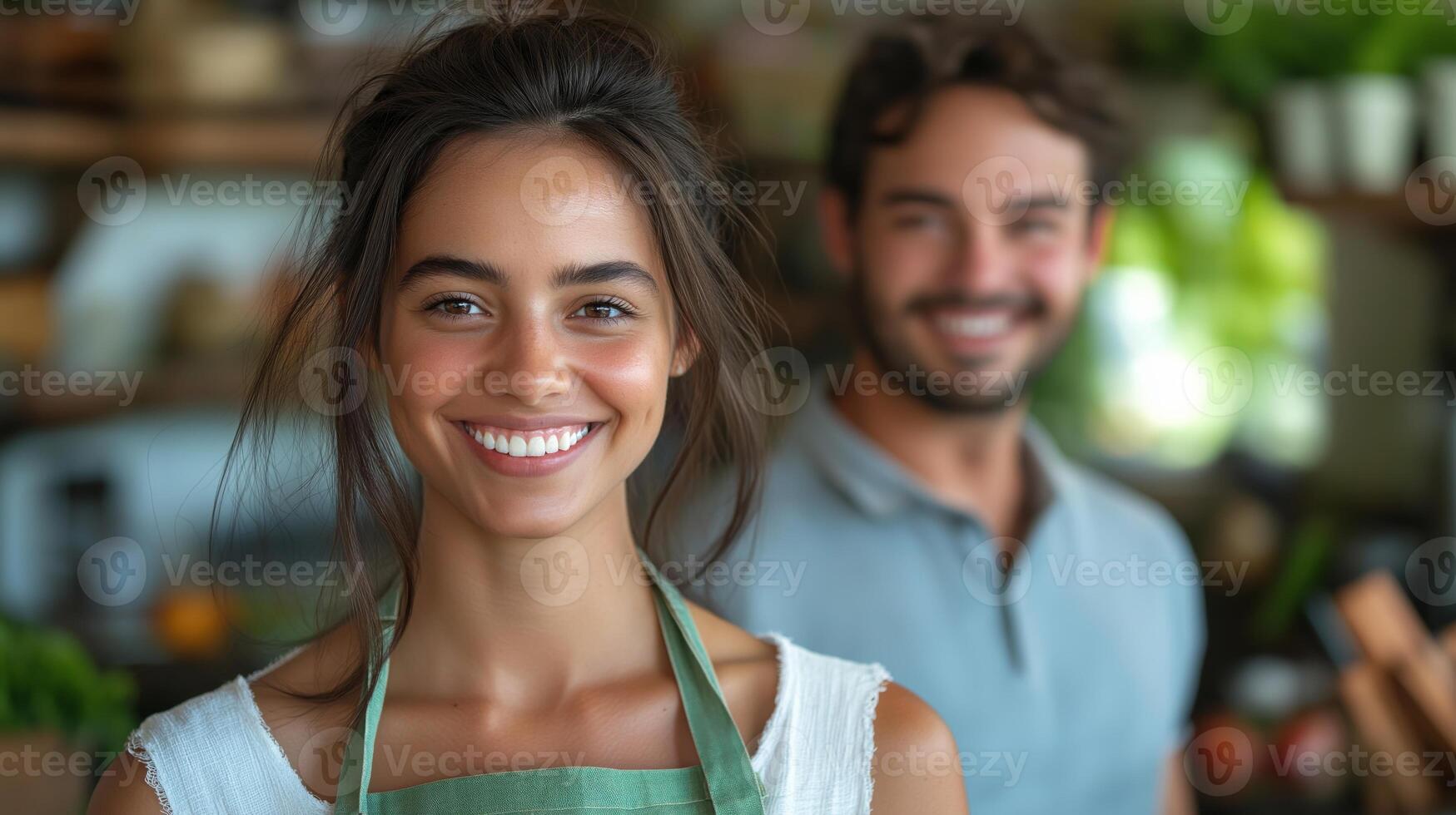 A young woman of mixed ethnicity smiles brightly while wearing an apron, with a supportive man in the background. photo