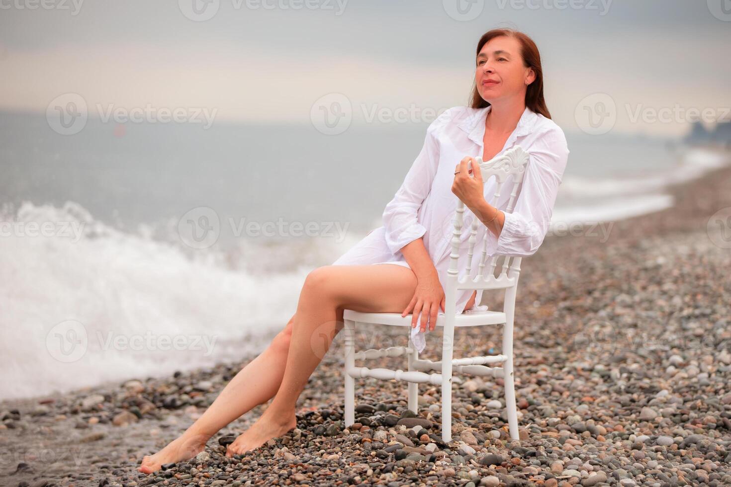 A woman is sitting on a chair on a beach. She is wearing a white shirt and shorts. photo