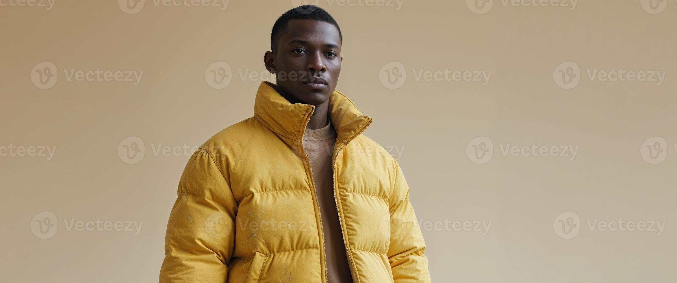A man wearing a yellow puffer jacket stands in front of a beige background. photo