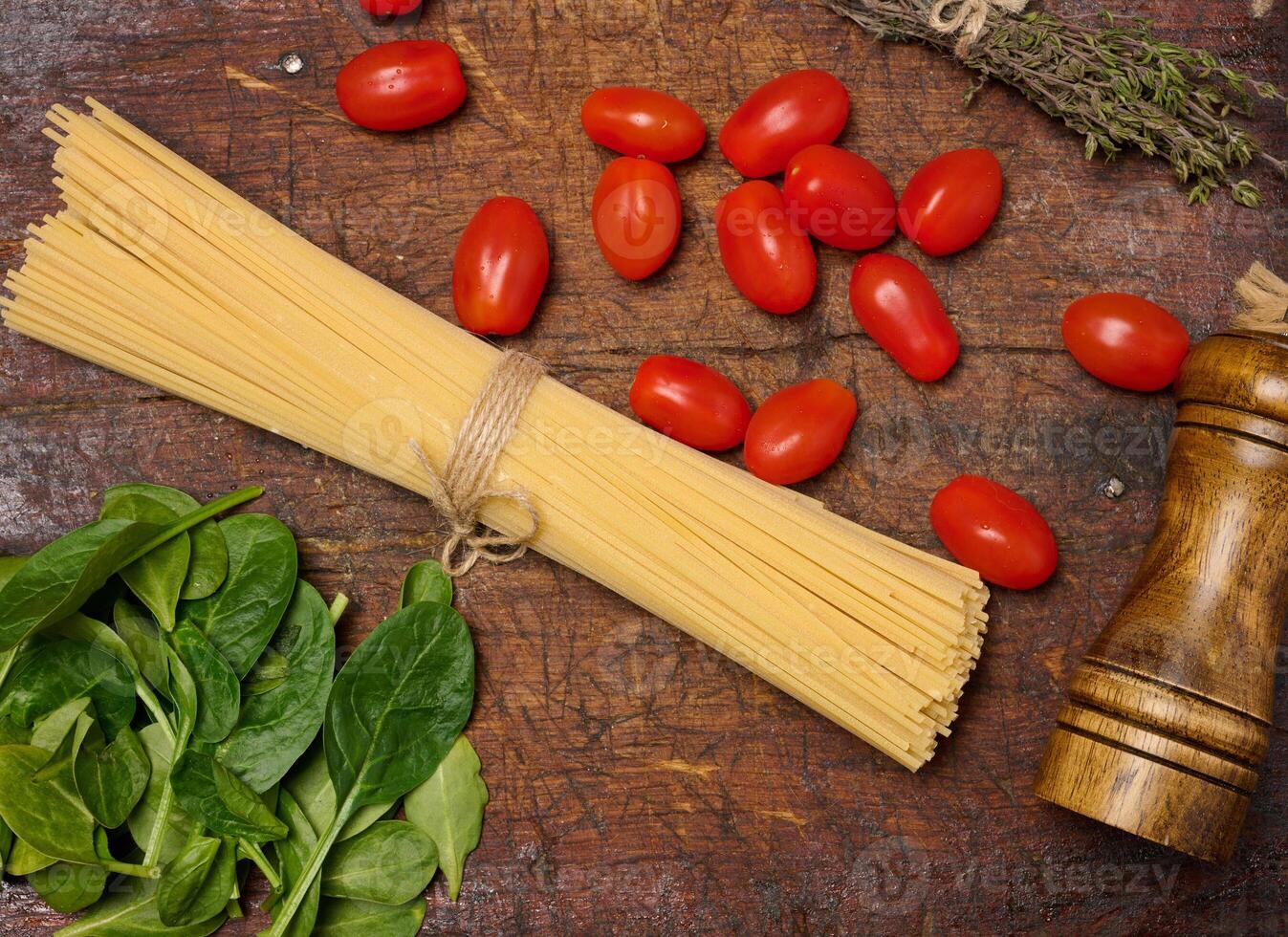 A bundle of raw pasta tied with twine, surrounded by cherry tomatoes, fresh spinach leaves, thyme, and a wooden pepper grinder on a rustic wooden surface. photo