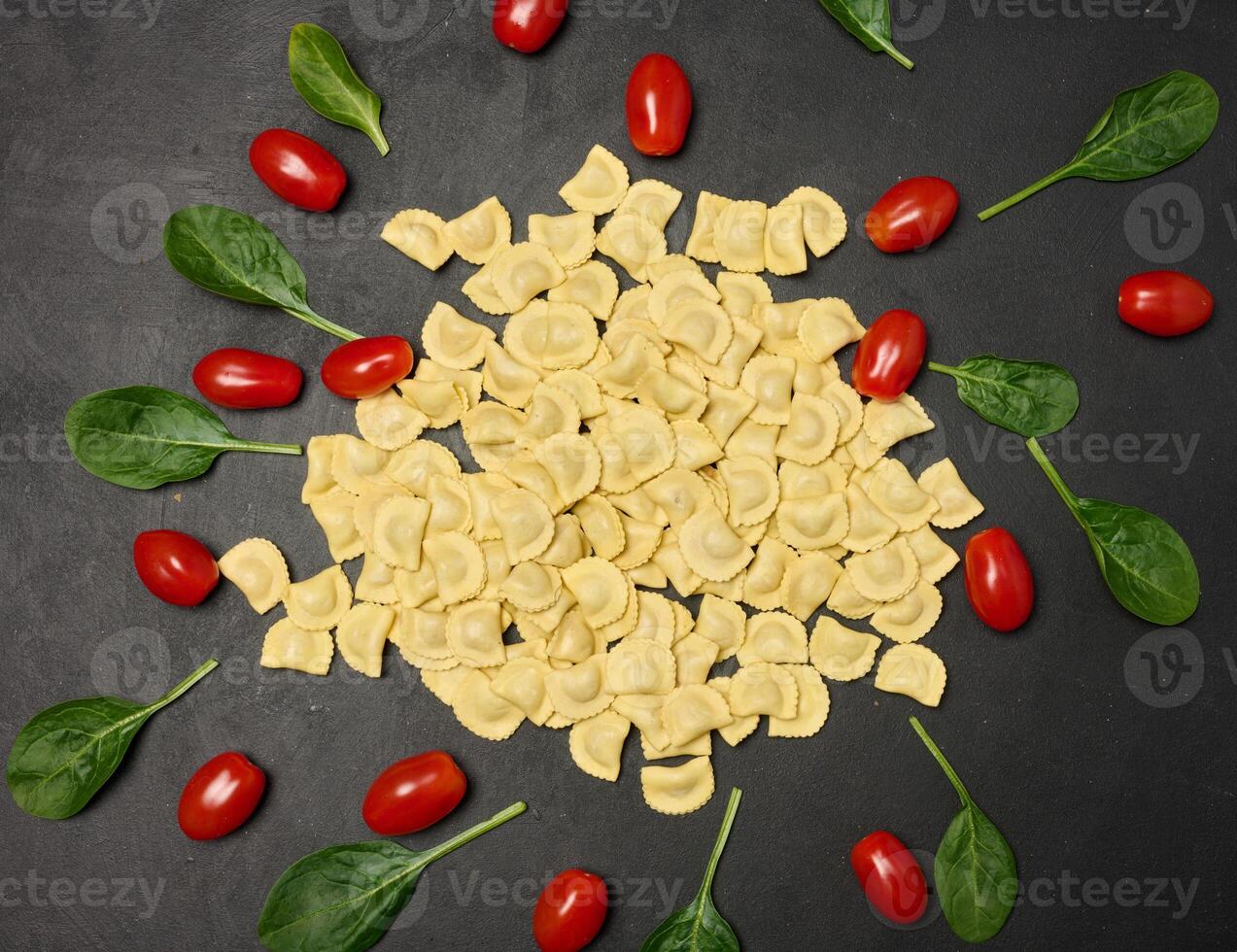 Uncooked tortellini pasta with fresh cherry tomatoes and spinach leaves on a dark, textured surface. Flat lay shot of ingredients arranged in a scattered pattern photo