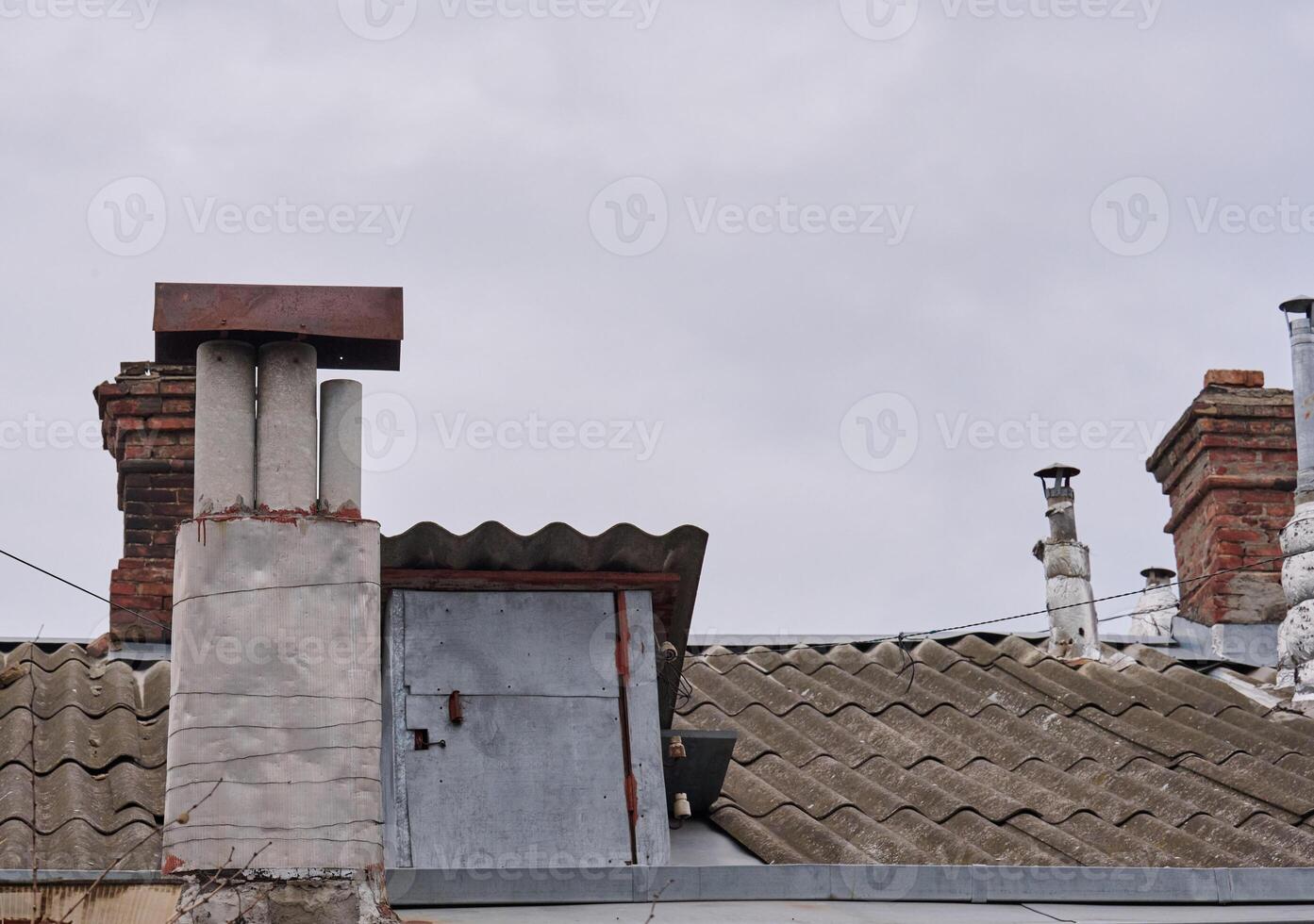 A close-up shot of several weathered chimneys on a rooftop, vary in design and material, some made of brick, others of concrete or metal, showcasing signs of age and exposure to the elements. photo