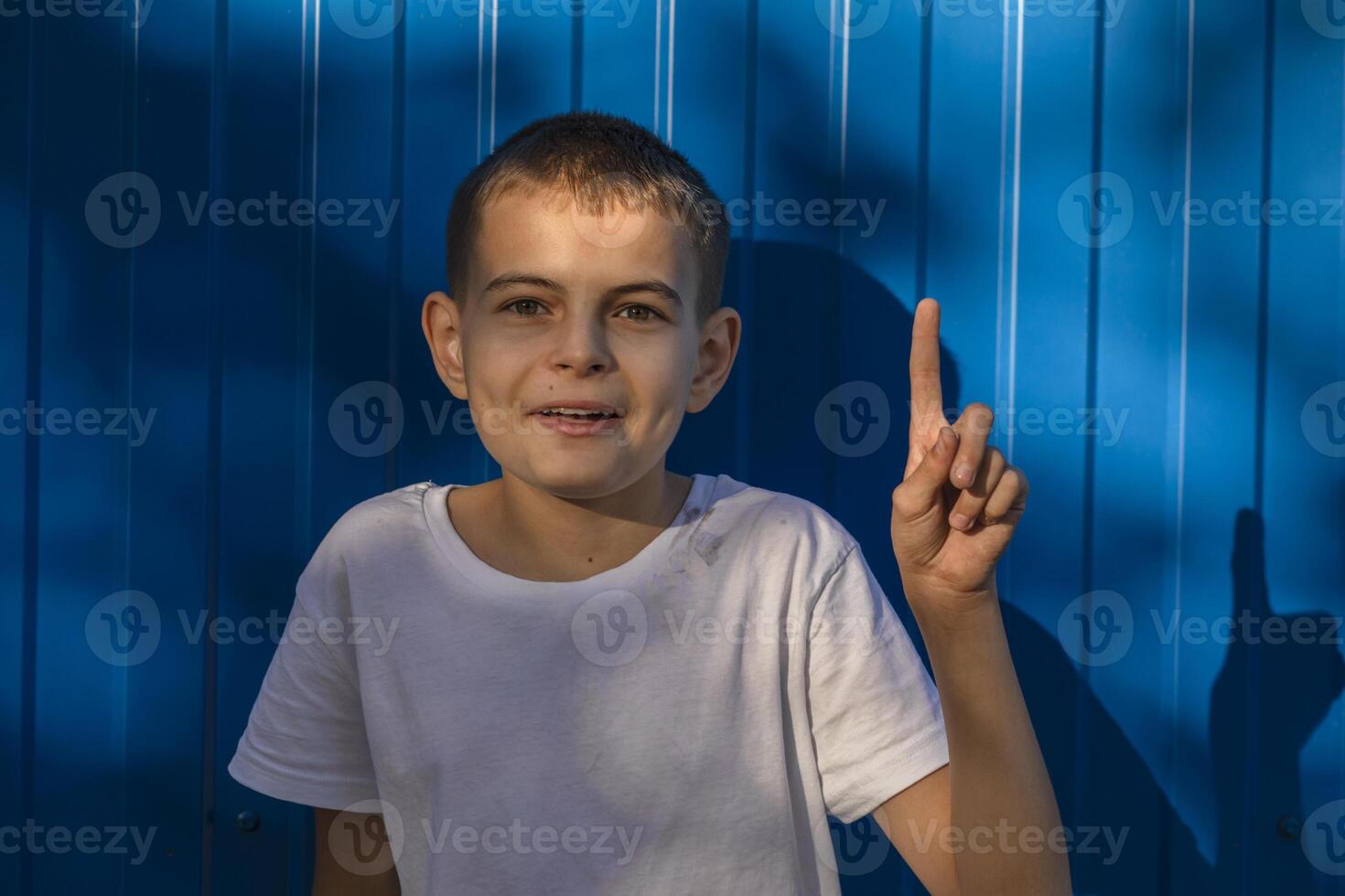 Little small happy boy 10 years old in whhite casual t-shirt point index finger aside on workspace area on plain blue background photo