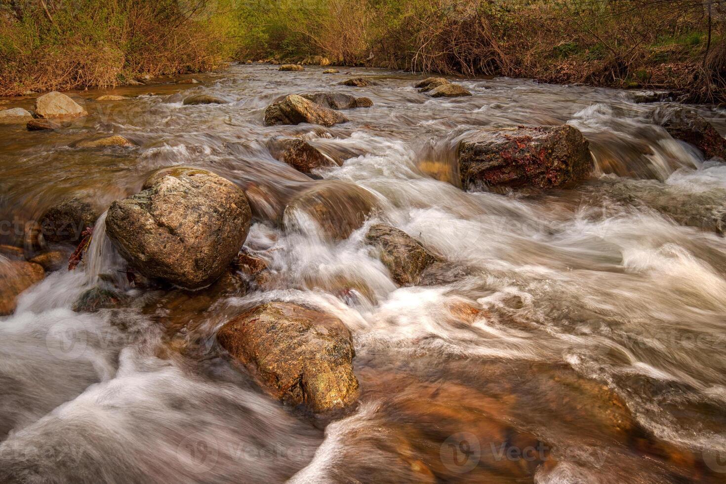 The water flows over numerous large, rounded rocks, creating a sense of movement and energy. photo