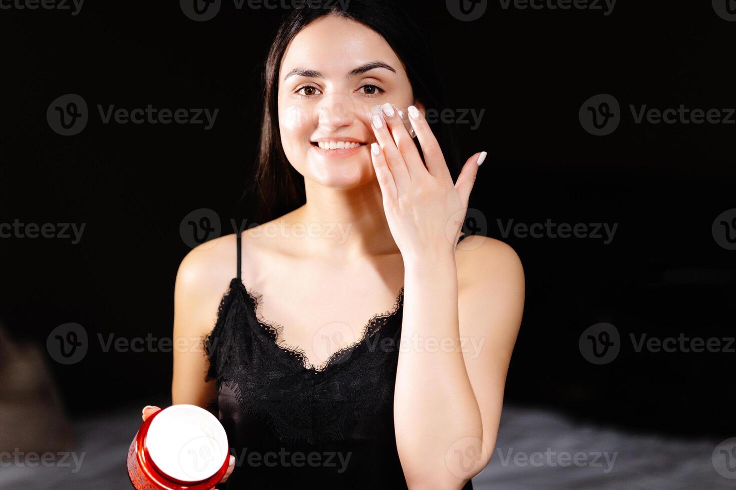 Young adult woman apply facial cream look at camera. Beautiful lady putting moisturizing lifting nourishing day cream on soft hydrated skin in bedroom. photo