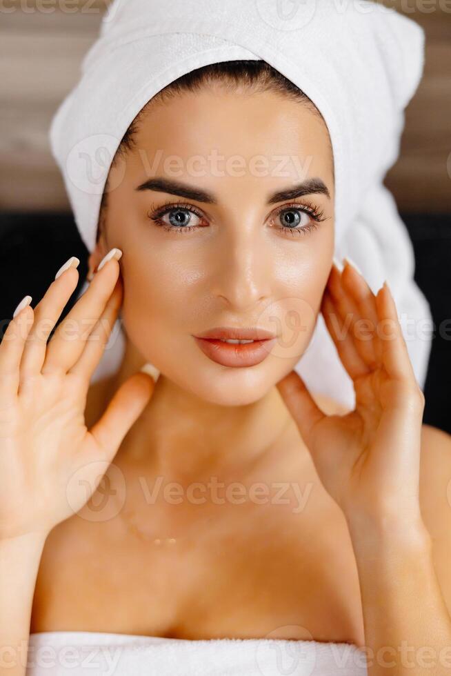 Happy beautiful woman wearing bathrobe and white towel with perfect complexion touching face looking at camera, sitting in room. Advertising of skin care spa wellness concept. Close up portrait. photo