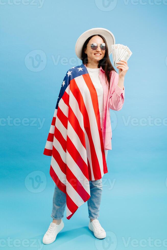 Young brunette woman in pink shirt poses on blue background in studio with american flag and hundred dollar cash. The smell of money photo