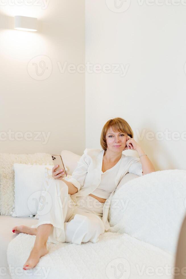 Beautiful woman relax siting on sofa at home and using cellphone texting chatting with friend. Happy young woman on couch holding smartphone in her hands, shoping online or check social media. photo