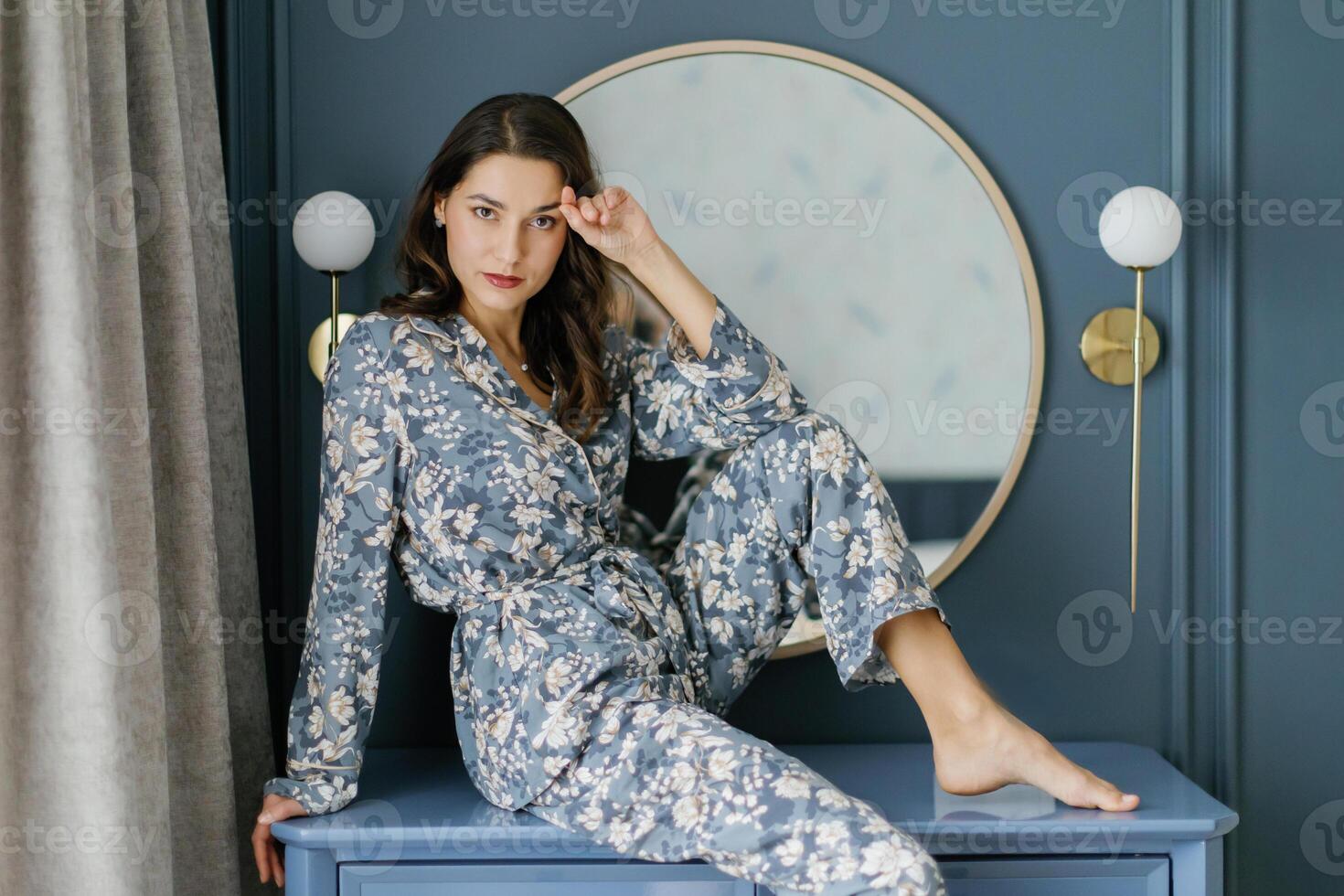 Woman wearing pajamas sitting on furniture in bedroom with round mirror. photo
