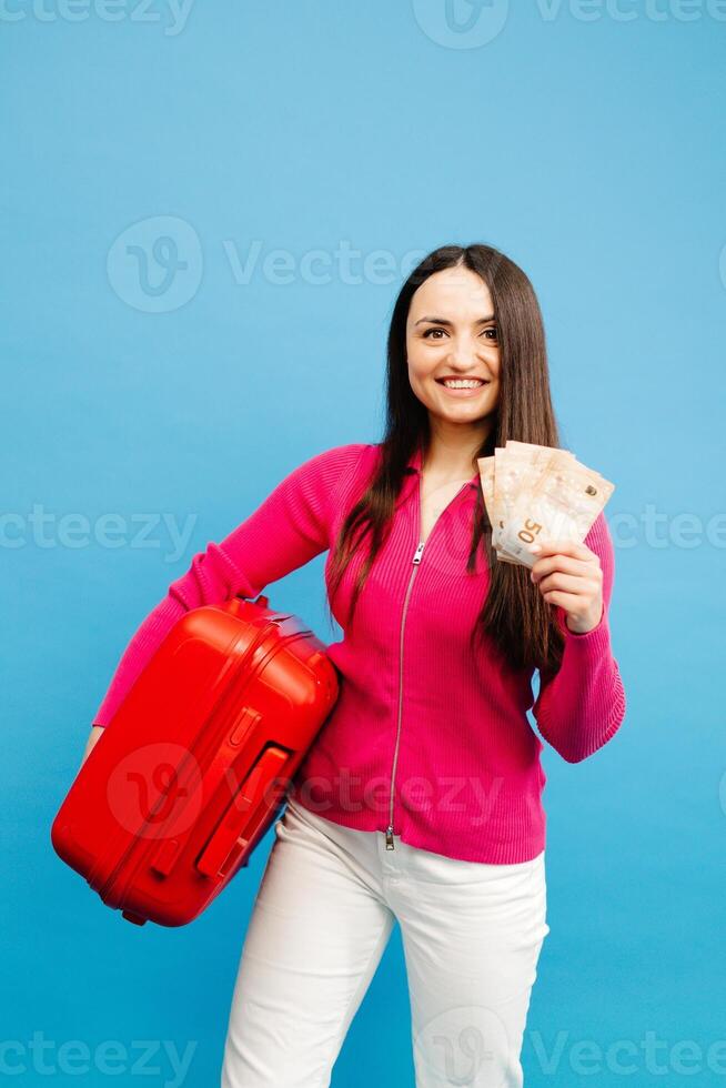 Positive girl with baggage and money are going to travel. Woman in pink sweater and jeans standing on blue background. photo