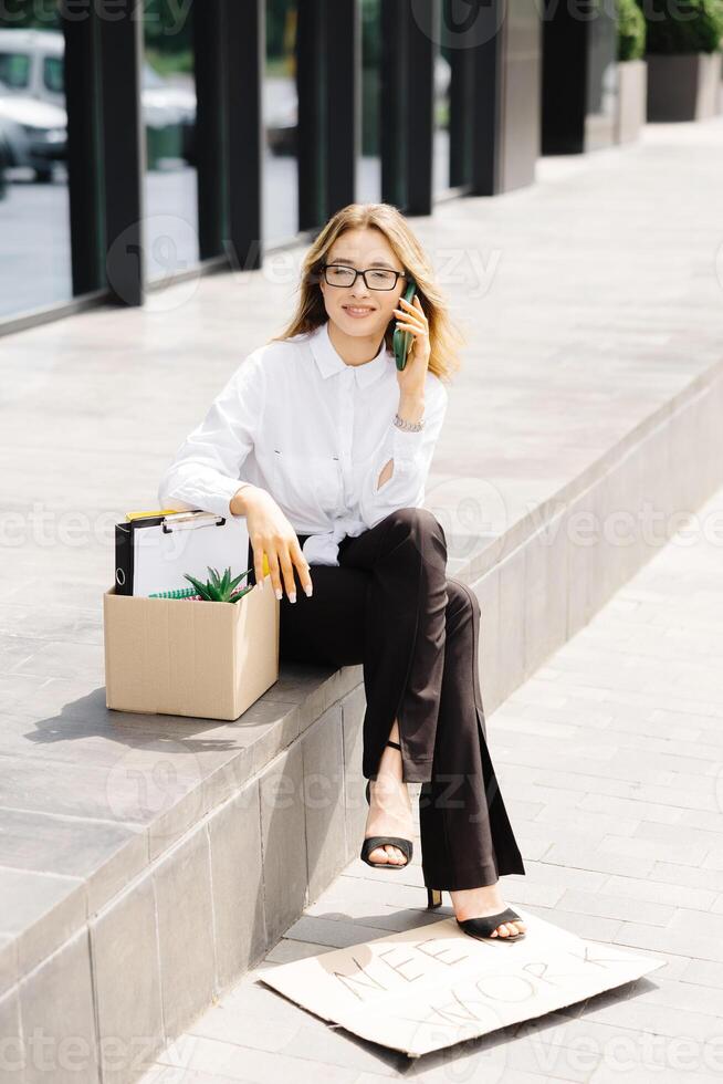Woman sitting outdoor with cardboard banner with the slogan Need work next to box of stuff and talking to the phone. Beautiful female protester at strike against unemployment photo