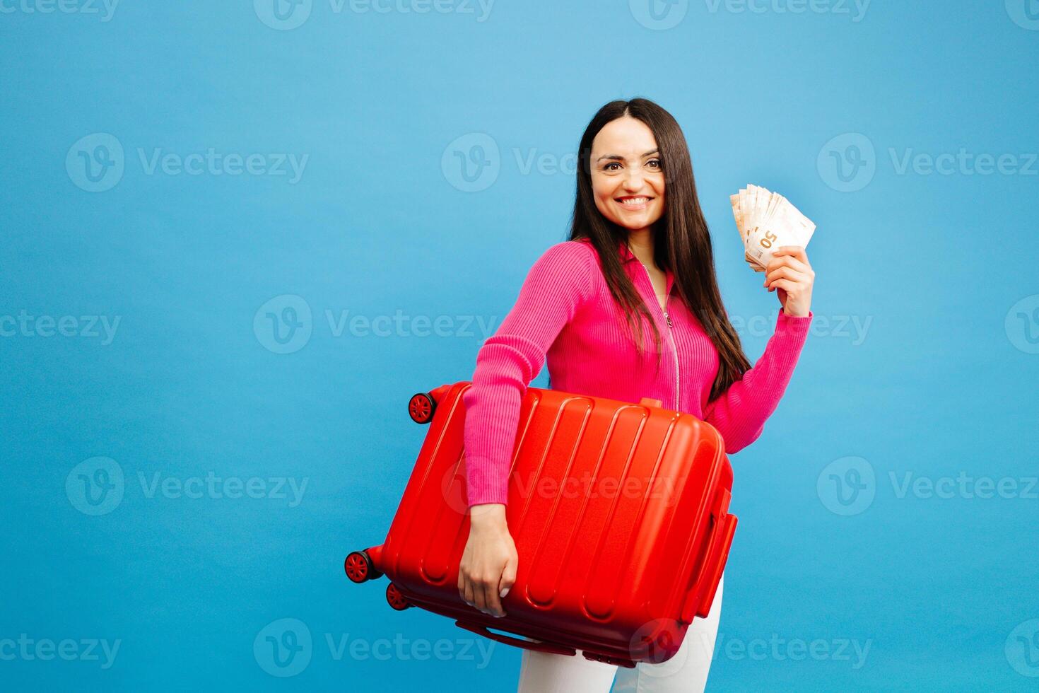Positive girl with baggage and money are going to travel. Woman in pink sweater and jeans standing on blue background photo