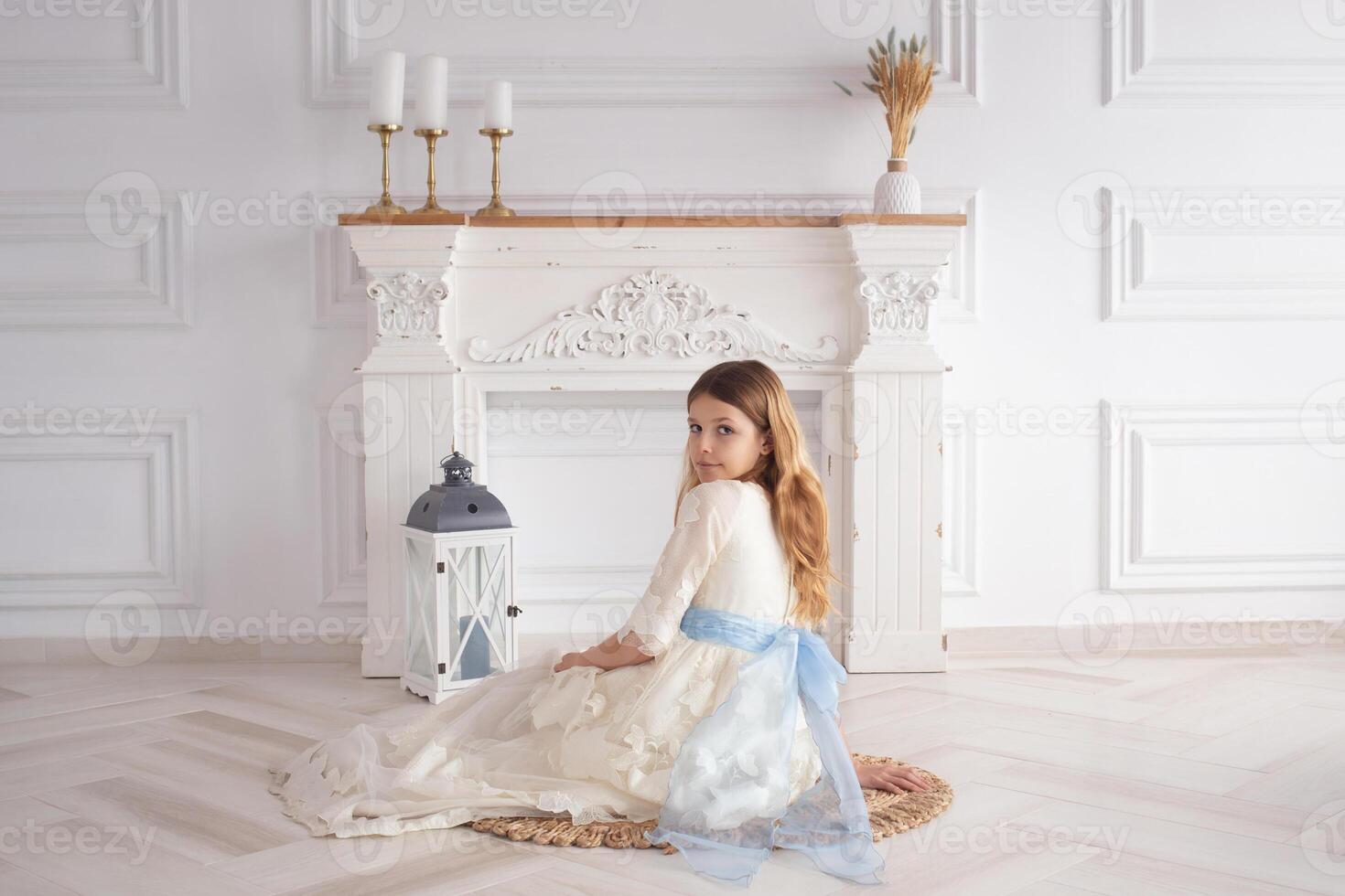 A beautiful girl in a smart white dress sits on the floor near the fireplace with candles on the day of her first communion photo