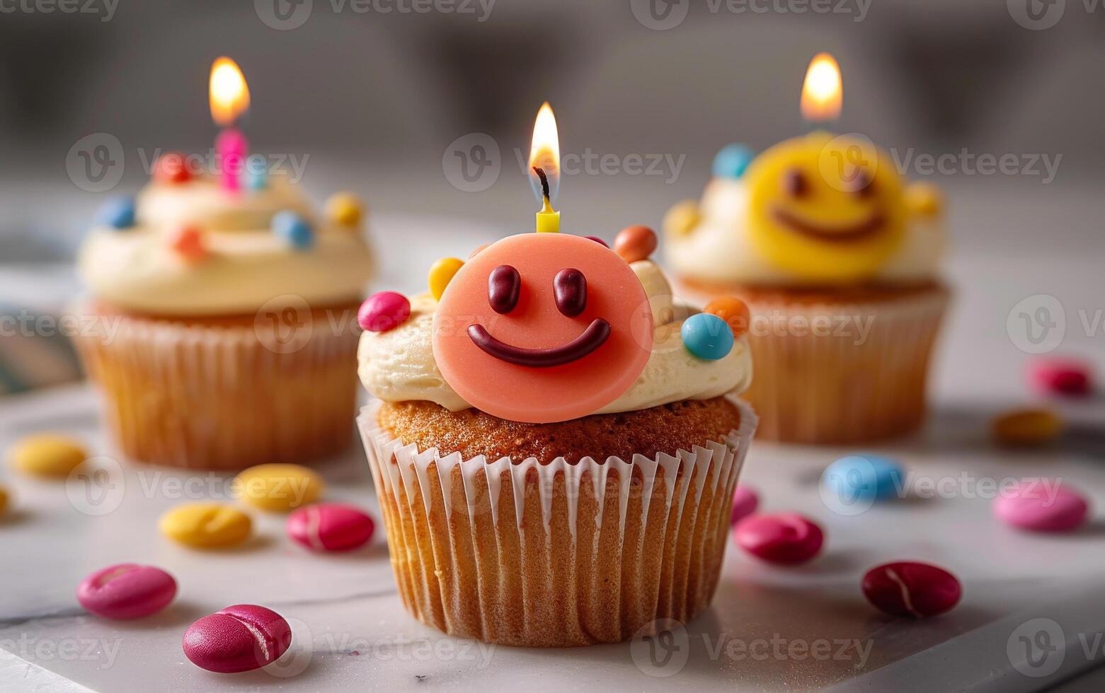 Smiley face cupcakes with candles. Three cheerful cupcakes with frosting and smiley faces are artfully arranged on a marble surface with candies. photo