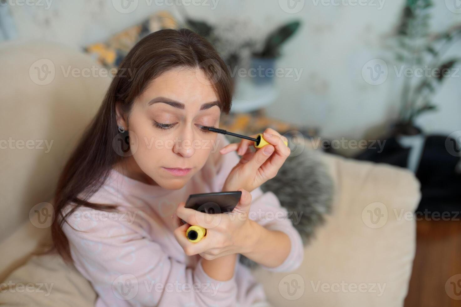 Young woman applying mascara, into handheld mirror while relaxing on living room sofa with soft background lighting morning routine photo