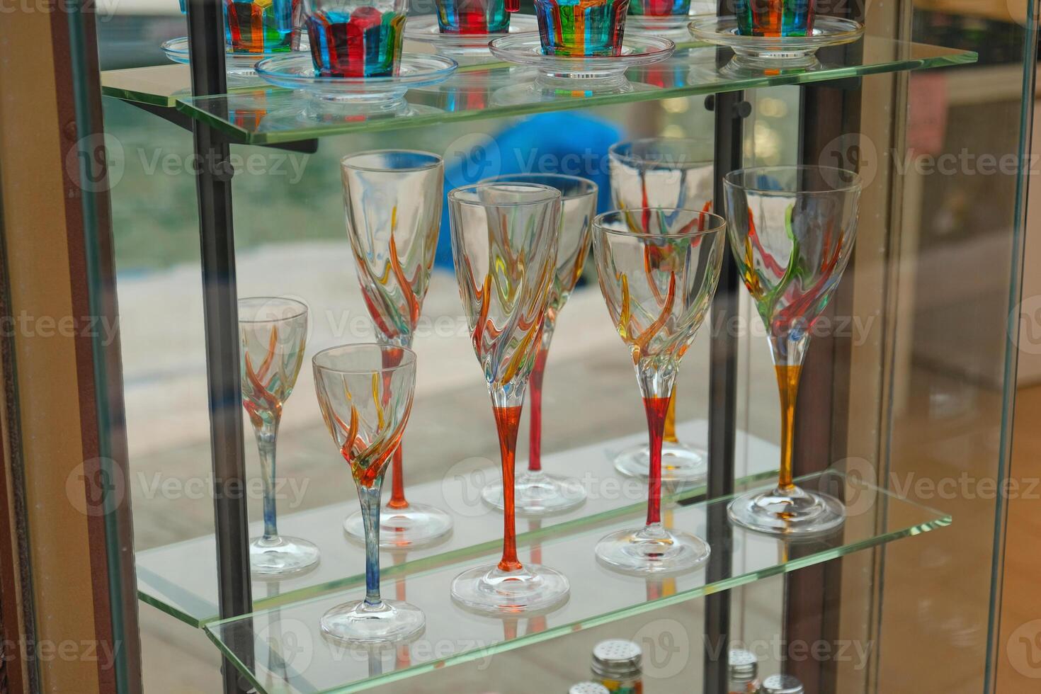 Elegant Murano glassware in Venetian style displayed on glass shelves in showcase store. Handcrafted artistic wine glasses and tumblers with vibrant multicolored designs, reflecting light. photo