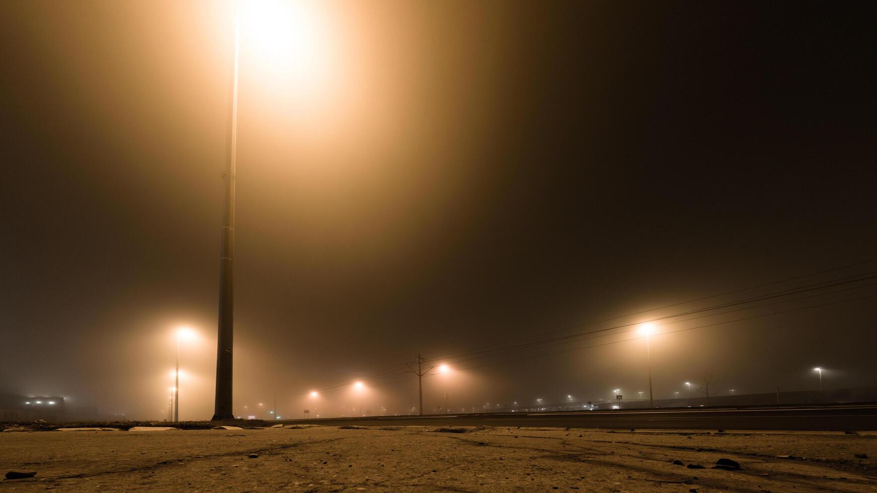 Streetlights cast an eerie glow through dense fog on a quiet, empty road at night, creating a moody and cinematic urban atmosphere. photo