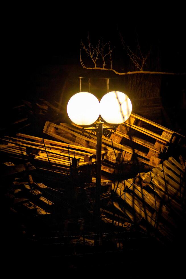 A glowing vintage street lamp stands against a dark backdrop, casting light over scattered wooden pallets. The eerie contrast between warmth and decay creates a mysterious, cinematic atmosphere. photo