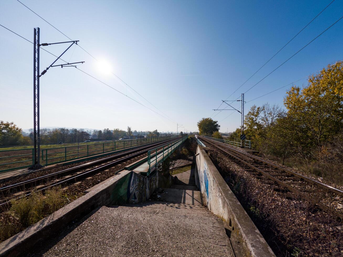 A pair of railway tracks extending into the distance under a bright blue sky, surrounded by autumn foliage and open fields, evoking a sense of travel and adventure. photo