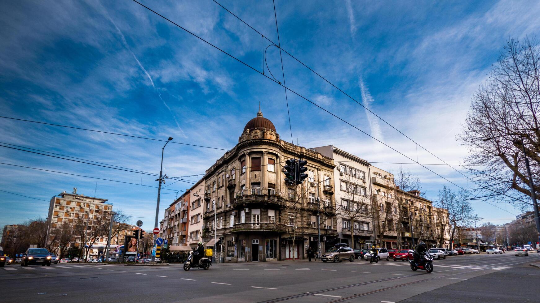 A grand, aged building with a domed rooftop stands at a lively urban crossroads, surrounded by modern city life, traffic, and tram wires under a vibrant blue sky. photo