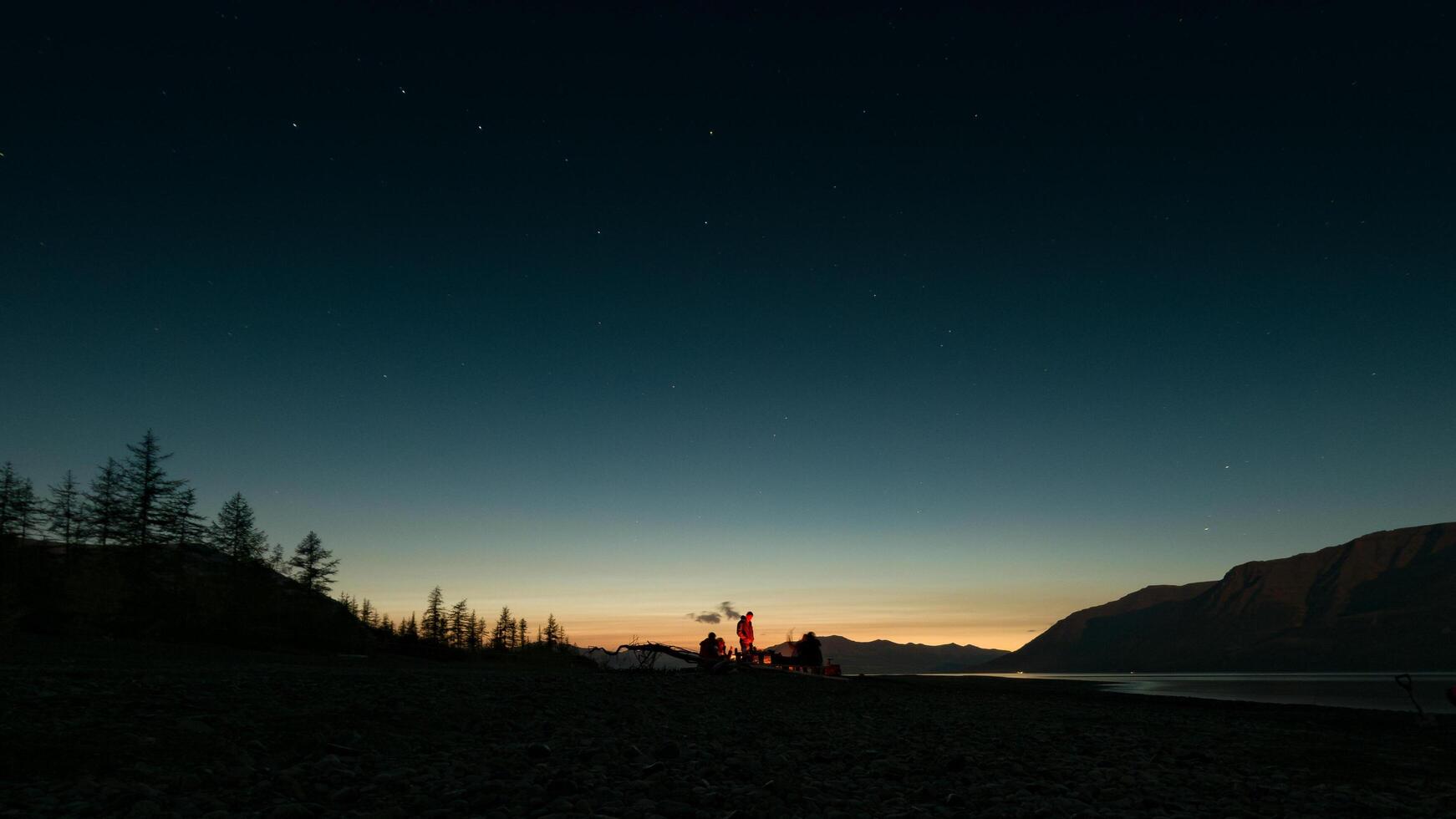 A serene nighttime scene featuring a group of people gathered around a campfire on a rocky lakeshore. The sky transitions from deep twilight blue to warm golden hues at the horizon. photo
