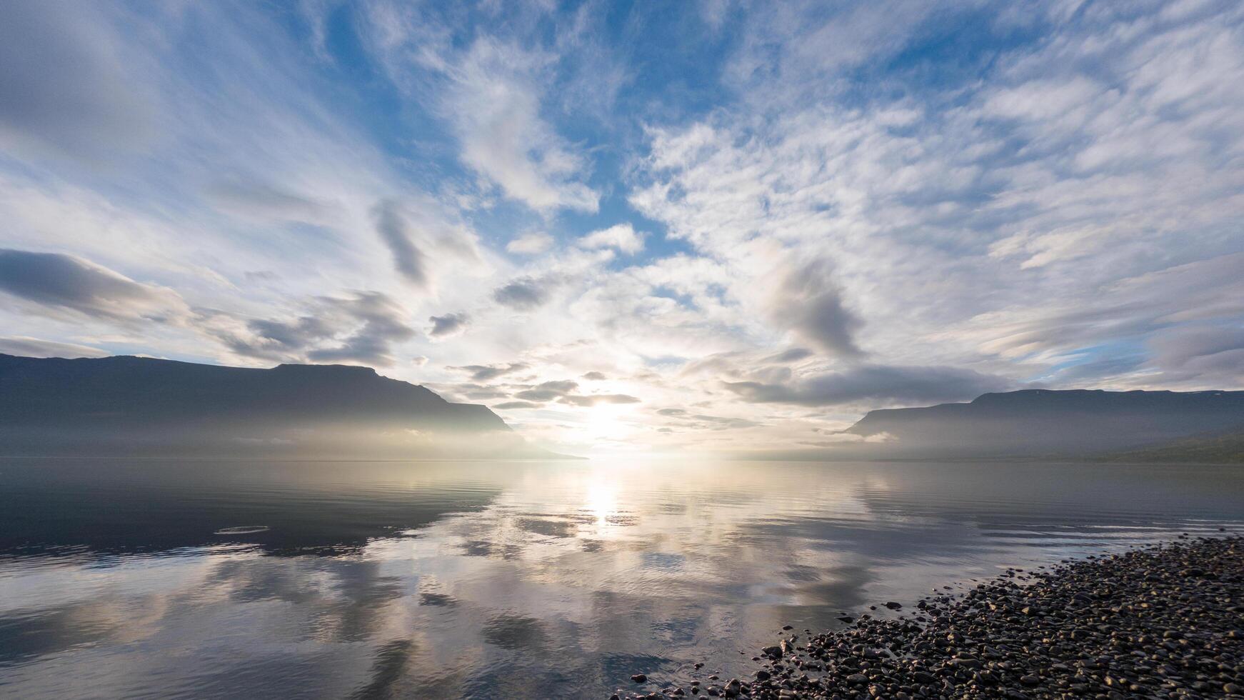The first light of dawn emerges from behind distant mountains, casting a golden glow across the tranquil lake. Wisps of mist hover gently above the water. photo