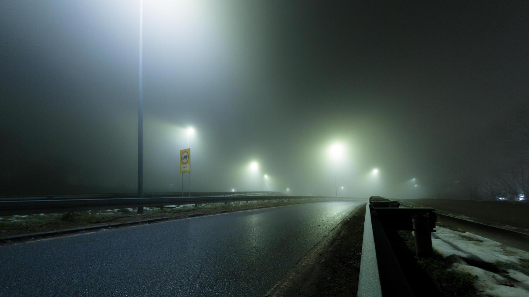 A desolate highway shrouded in thick fog, with glowing streetlights casting an eerie atmosphere. The misty road disappears into the unknown, evoking mystery and solitude. photo