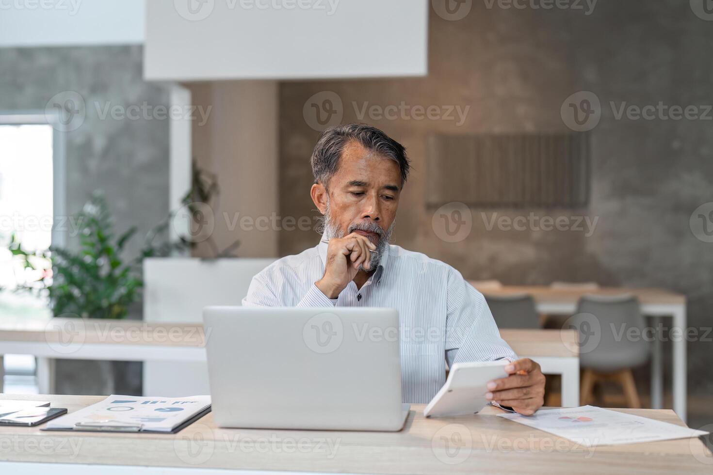 Elderly businessman thoughtfully calculating expenses with tablet and laptop in modern workspace photo