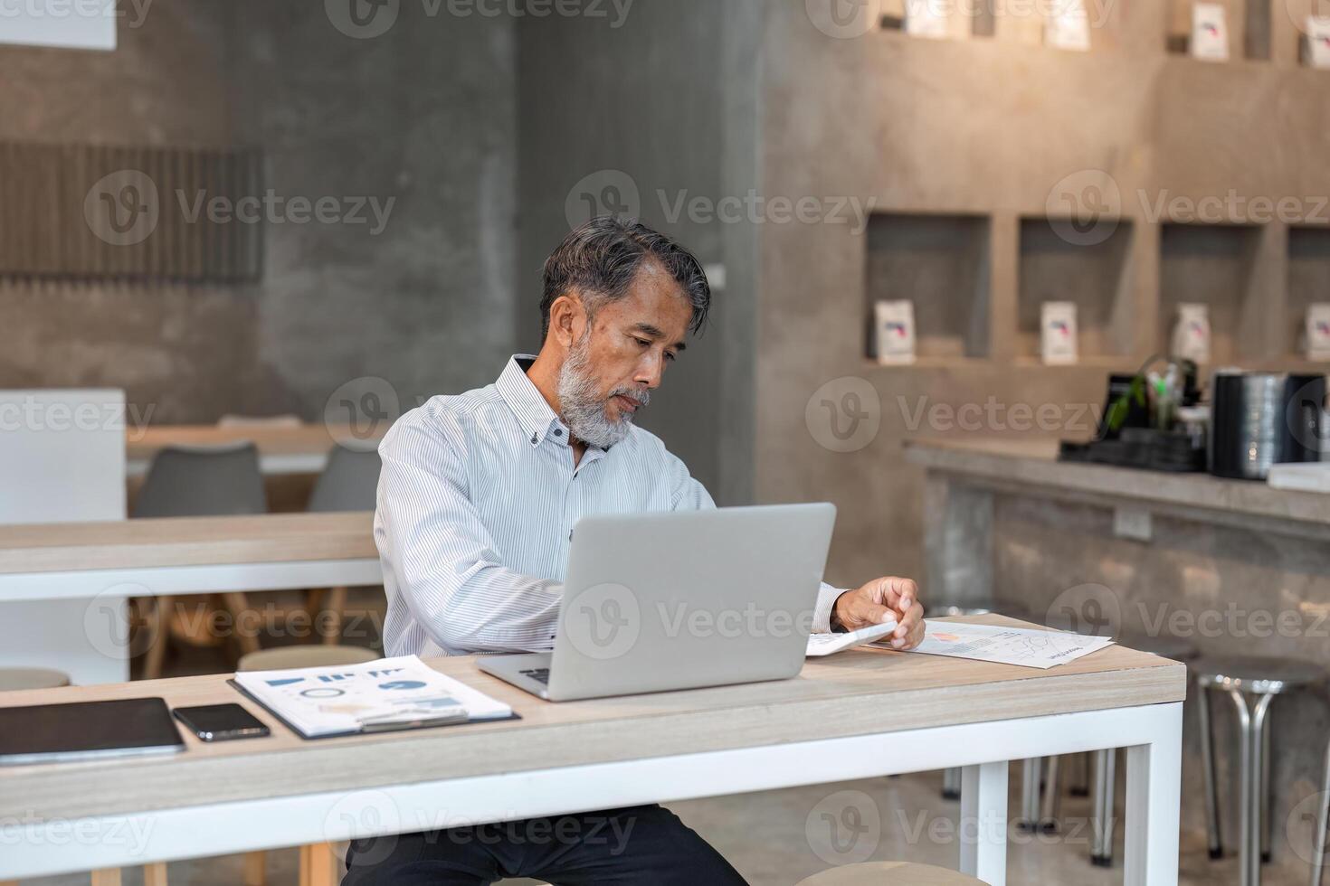 Elderly businessman analyzing financial documents while working on laptop in modern office photo