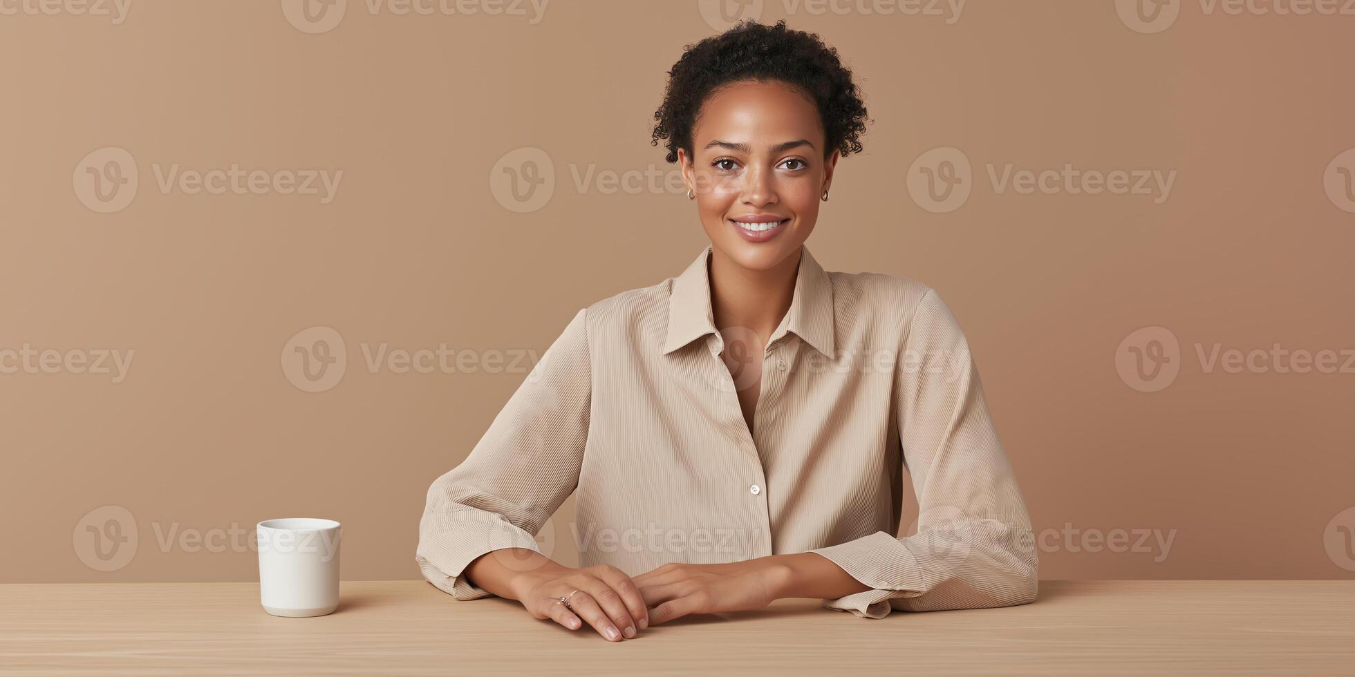 A confident person sitting at a table with a cup, wearing a beige shirt, smiling warmly. The background is a plain, neutral color, adding a sense of calm and focus. photo